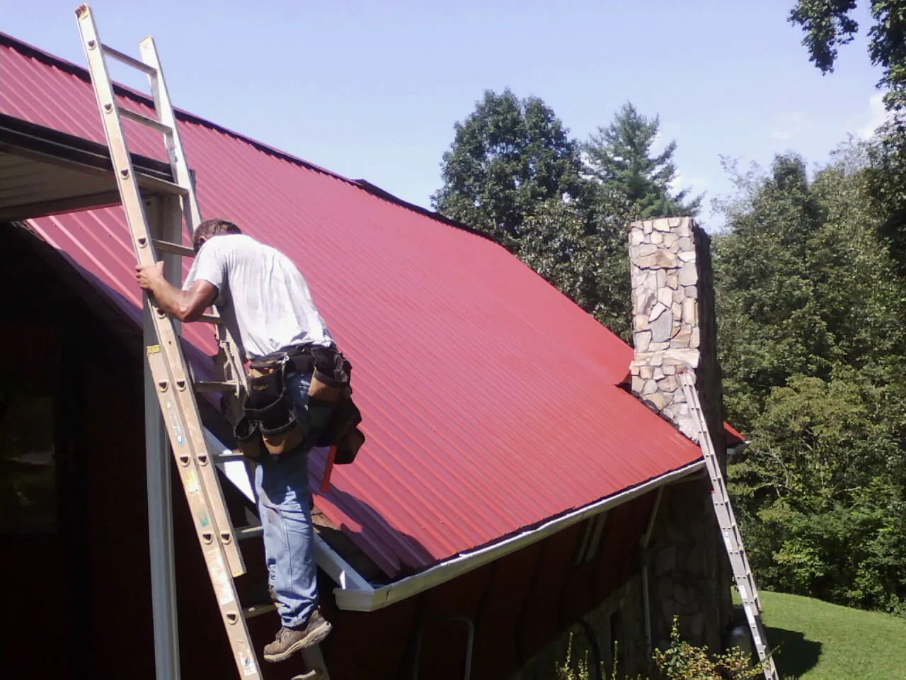 Roofer on a ladder beside a red metal roof and stone chimney — Soffit & Fascia Repair by Roofing Brothers in Hornbeak
